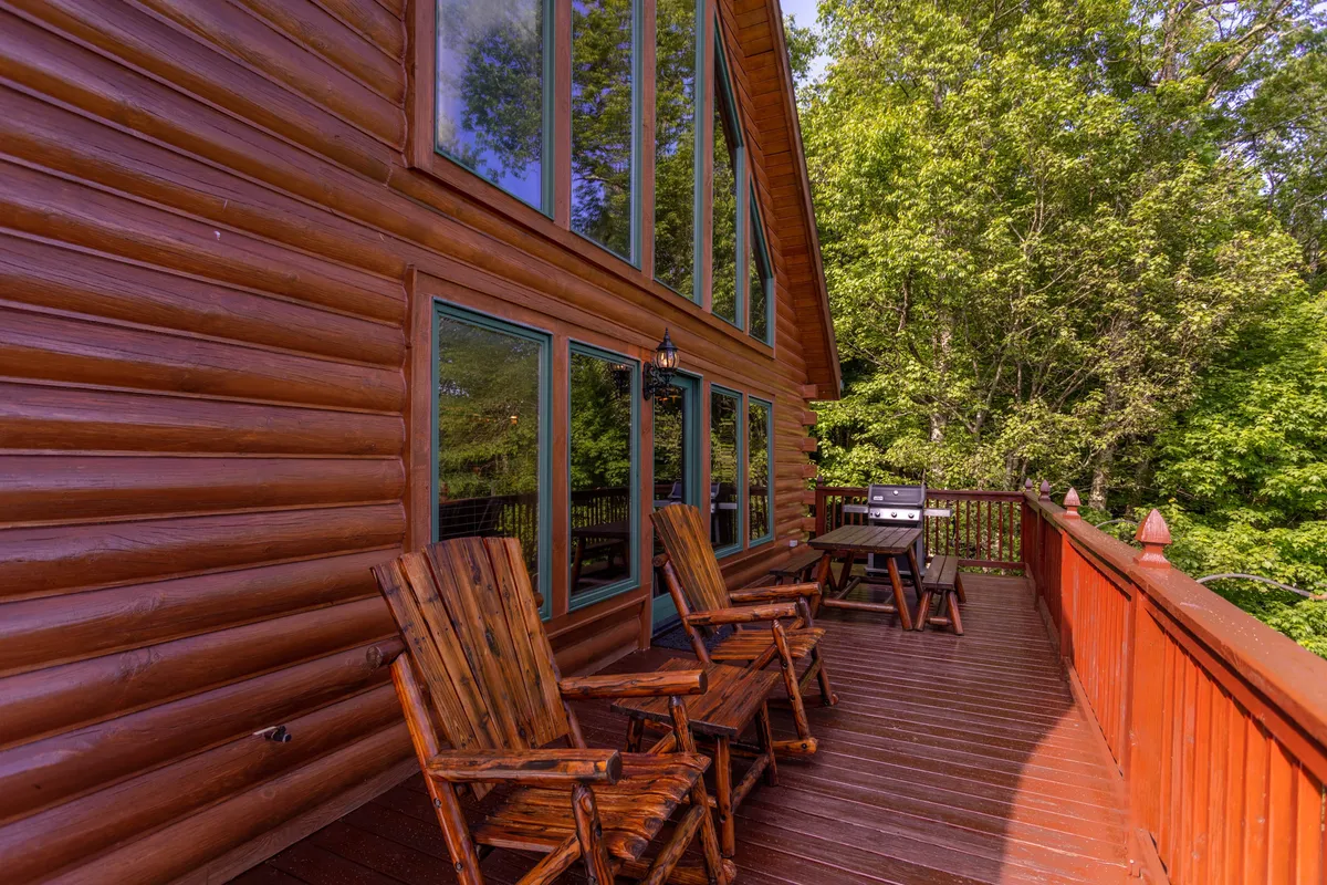 The photo provides a closer look at the deck’s amenities, including wooden chairs and a gas BBQ grill set against a backdrop of large windows and deck railing. The warm light suggests a setting perfect for evening barbecues or morning coffee with panoramic views.