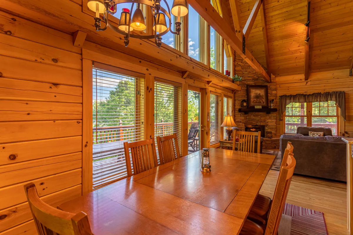 A dining area with a large table placed beneath wooden beams and a striking chandelier, framed by expansive windows that reveal natural light and scenic views. The setup is ideal for family meals, games, and celebrations.