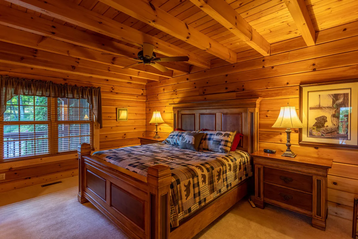A warmly lit bedroom with a plush bed, nightstands, a window that frames views of nature, and rustic ceiling beams. The atmosphere is peaceful and inviting, with cozy bedding and a tranquil setup for relaxation or reading.