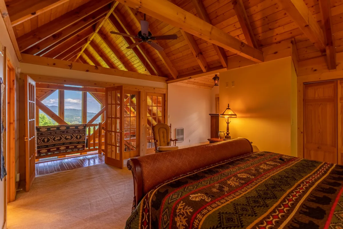 This image features a cozy bedroom with large windows that offer scenic views of the surrounding outdoors. The wooden ceiling beams and natural textures emphasize a connection to nature, while the comfortable chair positioned to enjoy the view encourages guests to engage with the landscape visible from indoors.