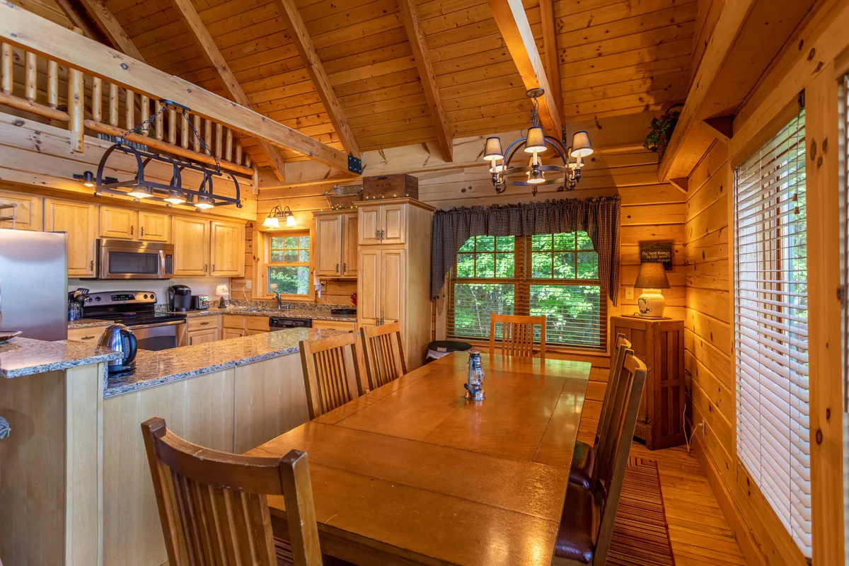 The photo highlights a large dining table set within an open kitchen layout, accented by wooden beams and natural light streaming through a window. The space is ready for shared meals, cooking together, and hosting memorable gatherings with ample seating for a group.