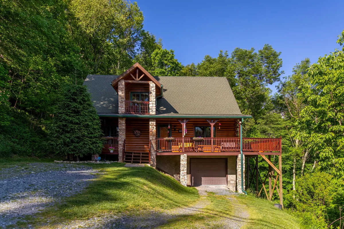 The image showcases a charming log cabin perched on a lush, green hillside in Banner Elk, North Carolina. The spacious wooden deck wraps around the cabin, offering panoramic views of the surrounding trees and distant Grandfather Mountain. Stone accents add rustic elegance, while the serene setting invites relaxation and outdoor enjoyment.