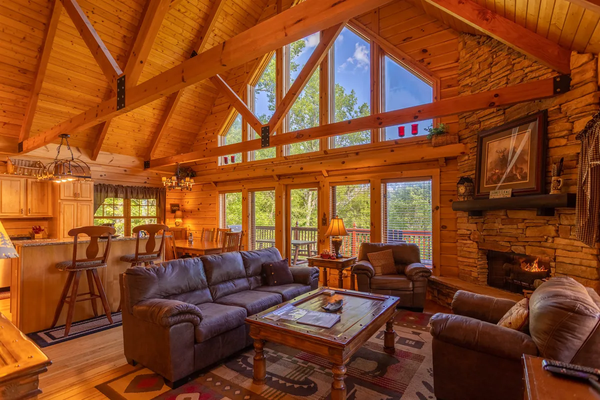 This image captures a warm, comfortable living area featuring large windows that frame the surrounding nature, a striking stone fireplace, exposed wooden beams, and rustic furniture. The scene depicts an inviting atmosphere perfect for family gatherings, cozy evenings by the fire, and relaxing after outdoor adventures.