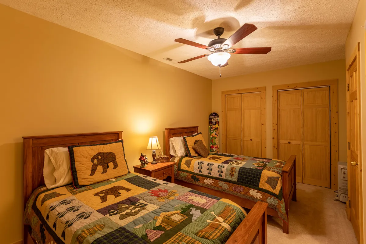 A cozy bedroom with two wooden twin beds covered in colorful quilts, a shared nightstand with a lamp, and a ceiling fan overhead. The scene suggests a welcoming, family-friendly space for kids or friends to rest and connect.