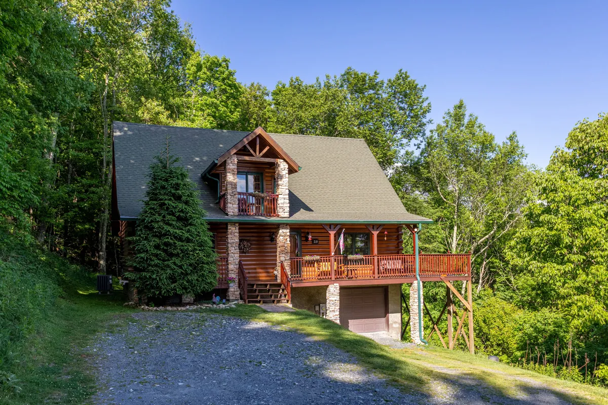 A broad view of the cabin's log exterior with stone accents, a large front porch, and lush greenery surrounding the property. The image captures the cabin's natural setting, emphasizing immediate access to outdoor adventures and the tranquil environment.