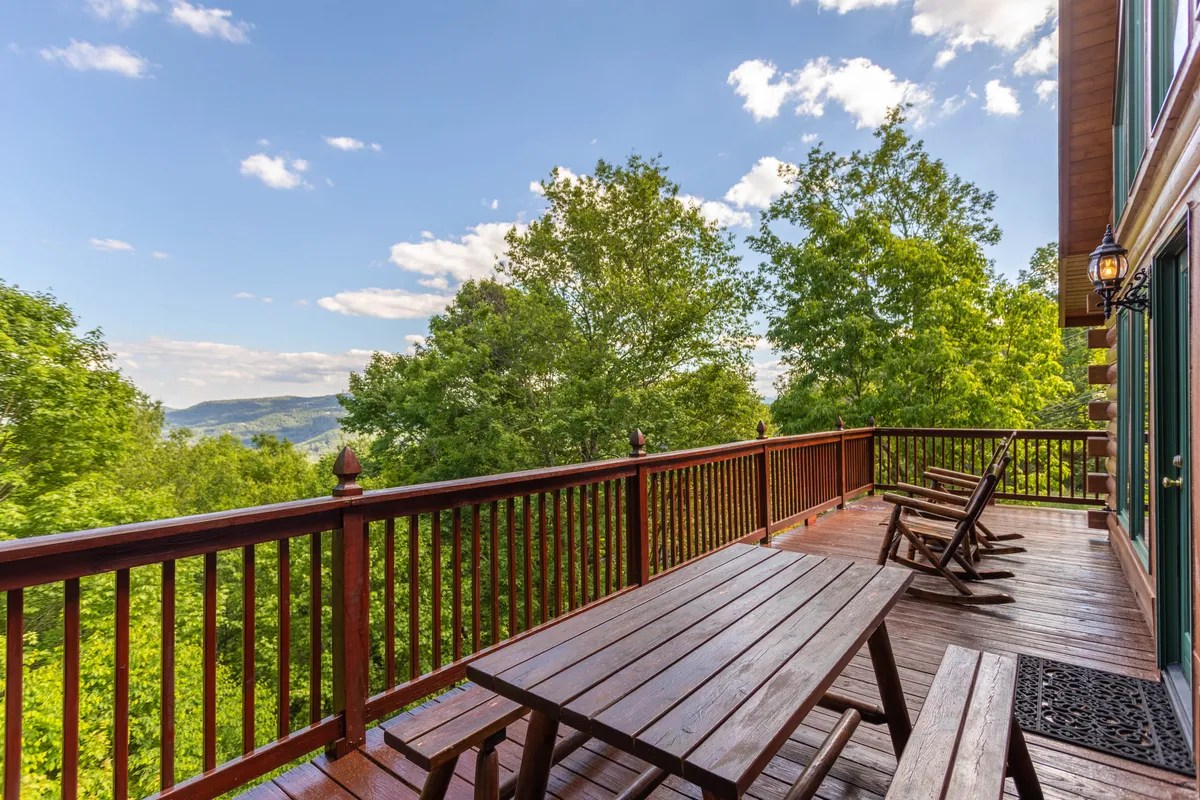 This image showcases a spacious wooden deck surrounded by lush greenery and overlooking mountain vistas. The deck features a log dining table perfect for alfresco meals, as well as Adirondack-style rocking chairs arranged for relaxation and conversation. The setting radiates tranquility, with ample space to gather, dine, or simply take in the natural beauty.